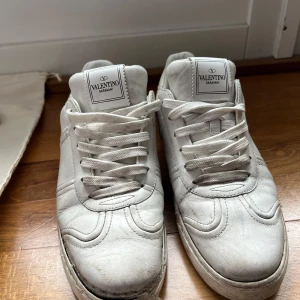 Vita Valentino Garavani sneakers - Säljer ett par vita sneakers från Valentino Garavani med klassisk design och diskreta nitar på sidan av sulan. Skorna har rund tå, snörning och är tillverkade i skinn. Perfekta för dig som gillar stilrena och exklusiva sneakers. Skorna har en skada som går att fixa. Du ser skadan på bild 5 sulan har låssnat men det går att lämna in hos en skomakare. Skriv om intresserad, jag kan skicka fler bilder om önskas. Skorna är stl 39 1/2. Vågar inte säga om de passar 40/uppåt. 