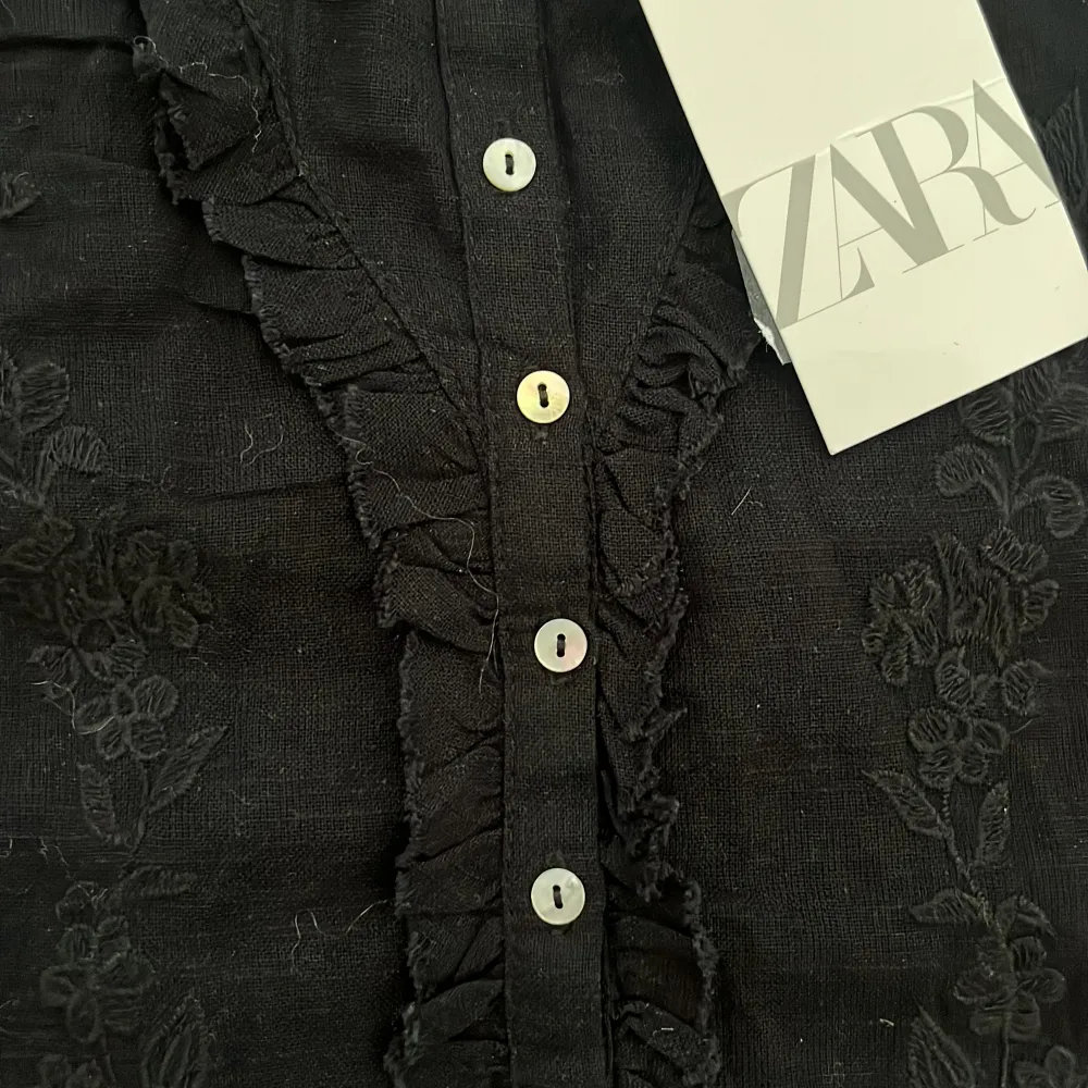 Snygg svart linnetopp från Zara med blommigt mönster och knappar framtill. Toppen har smala axelband som går att justera och volangdetaljer längs kanterna. Perfekt för en stilren look.. Yläosat.