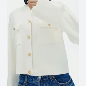 Vit cardigan jacka med guldknappar - Snygg vit cardigan jacka med dekorativa guldknappar och två bröstfickor. Jackan har en stilren design med långa ärmar och en rak passform. Perfekt för en elegant och avslappnad look.