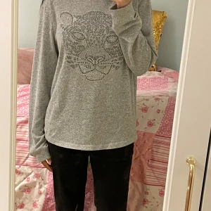 Grå hoodie med leopardmönster från Lindex - Säljer en grå hoodie från Lindex med ett stort leopardansikte i glittriga stenar framtill. Tröjan har långa ärmar och huva, perfekt för en avslappnad stil. Mjuk och skön bomullsblandning.