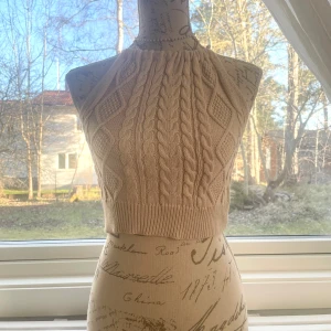 Beige stickad croppad halterneck-top - Säljer en beige stickad croppad halterneck-top med kabelstickat mönster. Toppen knyts i nacken och har ribbad nederkant. Perfekt för lager-på-lager eller som statement-piece till jeans eller kjol. Passar storlek XS-S.