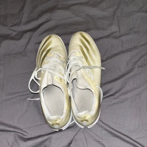 Vita fotbollsskor med guldiga detaljer - Säljer ett par vita fotbollsskor med guldiga detaljer och snörning. Skorna har en modern design med en rund tå och är perfekta för fotbollsplanen. Har ingen box med skorna.