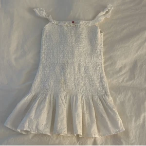 Vit kortklänning med volangkant - Supersöt vit kortklänning med smockad överdel och volang nertill. Axelbandslös modell med liten volang upptill och broderade detaljer på kjolen. Perfekt för sommarens alla tillfällen! Prislappen är kvar eftersom den är helt oanvänd 