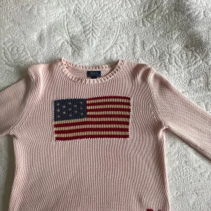 Ljusrosa stickad tröja från Polo Ralph Lauren - Säljer en ljusrosa stickad tröja från Polo Ralph Lauren med amerikansk flagga på bröstet och broderade initialer 'RL' nertill. Tröjan har rund hals och långa ärmar. Perfekt för dig som gillar american Clean girl style!