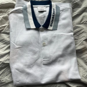 Vit pikétröja från Burberry - Säljer en stilren vit pikétröja från Burberry med klassisk krage i blå och grå toner samt märkets logga broderad på kragen. Tröjan har knäppning framtill med två knappar och är perfekt för en clean och snygg look.