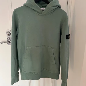 Stone Island hoddie - Säljer en stilren grön hoodie från Stone Island i Junior storlek 170 med ikonisk logga på ärmen. Passar bra till jeans. Köpt på NK i Stockholm för ca 2000 kr, säljer för 599 kr.
