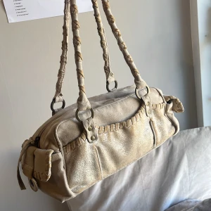 Beige axelväska med flätade detaljer - Snygg beige axelväska med unika flätade detaljer på handtagen och kanterna. Väskan har en praktisk dragkedja upptill och sidofickor med knapplås. Perfekt för att ge din outfit en bohemisk touch.