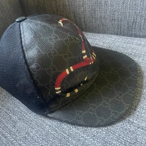 Svart keps med orm från Gucci - Snygg svart keps från Gucci med ett coolt ormmönster i rött och beige. Kepsen har ett diskret mönster av Gucci-loggor och är tillverkad i Italien. Perfekt för att ge din outfit en lyxig touch. Den är XS men passar 100% för er som har S då jag själv har S på mina andra Gucci kepsar 