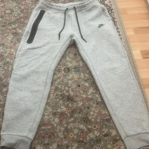 Grå mjukisbyxor från Nike - Säljer ett par grå mjukisbyxor från Nike med svart dragkedja på fickan och snörning i midjan. Perfekta för en avslappnad stil. Byxorna har en elastisk midja och muddar vid bensluten.