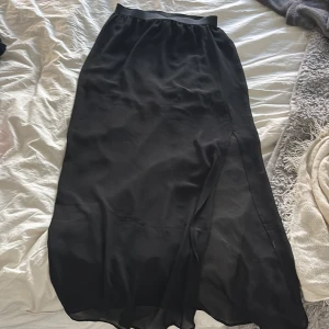Svart långkjol från H&M Divided - Svart långkjol från H&M Divided med elastisk midja och transparent nederdel. Kjolen har en slits på sidan och är tillverkad i lätt och luftig polyester, perfekt för en trendig look.