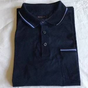 Mörkblå pikétröja från Abdullah Kiğılı - Snygg mörkblå pikétröja från Abdullah Kiğılı med kontrasterande vita och blå ränder på krage och ärmslut. Tröjan har en klassisk pikékrage med knappar och en liten bröstficka. Perfekt för en stilren look.