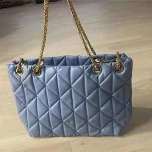 Blå quiltad axelväska med guldkedja - Snygg blå axelväska med quiltat mönster och guldfärgade kedjehandtag. Perfekt för att ge en touch av elegans till din outfit. Väskan har en stilren design som passar till många tillfällen.