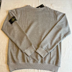Grå sweatshirt från Stone Island - Säljer en stilren grå sweatshirt från Stone Island. Tröjan har en rund halsringning och långa ärmar med ribbade muddar. Den ikoniska Stone Island-loggan är broderad på ärmen. Perfekt för en avslappnad och trendig look.