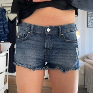 Mörkblå jeansshorts med fransar - Säljer ett par snygga mörkblå jeansshorts med fransiga kanter. De har en klassisk femficksdesign och en knappgylf. Perfekta för en avslappnad stil.