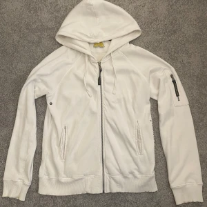 Vit hoodie från Acqua Limone - Säljer en vit hoodie från Acqua Limone med dragkedja och huva. Tröjan har två fickor fram och en broderad logga i gult och svart på ryggen. 