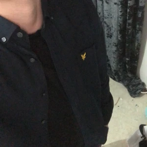 Svart skjorta från Lyle & Scott - Säljer en svart skjorta från Lyle & Scott med klassisk krage och knappar framtill. Skjortan har en gul broderad logga på bröstet och är långärmad. Perfekt för en stilren look.