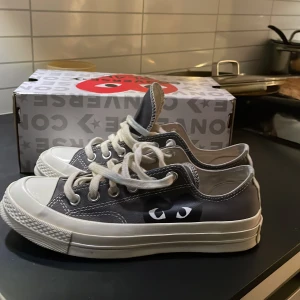 Converse Play x Comme des Garçons sneakers - Säljer ett par Converse Play x Comme des Garçons sneakers med det ikoniska hjärtmotivet på sidan. Skorna är grå med vita detaljer, vita snören och sula. Perfekta för dig som vill ha något unikt och trendigt i garderoben.