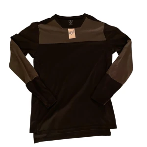 Fox tröja - Säljer en svart långärmad tröja från Fox med gråa paneler över axlar och armar. Tröjan har tryckt logga på axeln och vid halsringen. Perfekt för dig som cyklar. Ignorera längst ner på änden av tröjan (tröjan blev lite konstigt vikt).