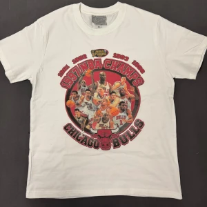 Vit Chicago Bulls t-shirt med NBA Champs tryck - Vit t-shirt med stort färgglatt tryck på framsidan som hyllar Chicago Bulls NBA-mästerskap 1991-1993. Motivet visar flera spelare och texten 'NBA Champs' samt 'Chicago Bulls' i rött och svart. Perfekt för basketfans!                                         Passar som L