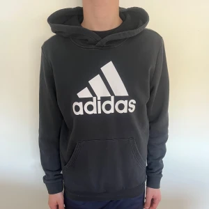 Svart hoodie från Adidas - Säljer en svart hoodie från Adidas med stor vit logga på bröstet. Tröjan har huva och en stor magficka framtill. Perfekt för en avslappnad och sportig stil.