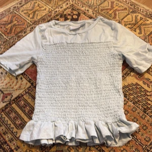 Ljusblå topp med volang från Gina Tricot - Säljer en ljusblå topp från Gina Tricot med smockat parti framtill och volang längst ner. Toppen har korta ärmar och rund halsringning. Perfekt för dig som gillar söta detaljer och en bekväm passform.