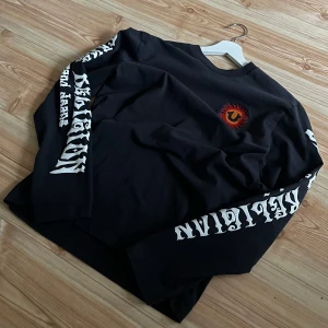  True Religion - Mörkgrå långärmad tröja från True Religion med tryck i vitt längs båda ärmarna och ett färgglatt logomärke på bröstet. Tröjan har en avslappnad passform och är perfekt för en streetwear-look.