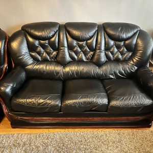 Svart soffgrupp i läder med trädetaljer - Säljer en klassisk soffgrupp i svart läder med snygga trädetaljer. Gruppen består av en tresitssoffa, en tvåsitssoffa och två fåtöljer. Snyggt mönstrad rygg och breda armstöd ger en lyxig känsla. Perfekt för vardagsrummet.
