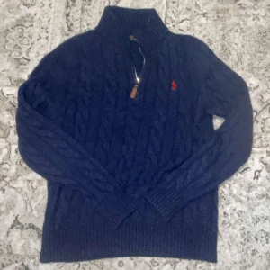 Mörkblå stickad tröja med dragkedja från Polo Ralph Lauren - Mörkblå stickad tröja från Polo Ralph Lauren med halv dragkedja och hög krage. Klassisk kabelstickad design och röd broderad logga på bröstet. Perfekt för lager-på-lager och stilren look. Jätteskön till sena vårkvällar skulle jag säga, helt nya oanvänd.