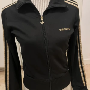 Svart träningsjacka från Adidas med guldiga detaljer - Snygg svart träningsjacka från Adidas med guldiga ränder längs ärmarna och guldigt Adidas-tryck på bröstet. Stolkek är 38 men skulle säga med 36/34. 
