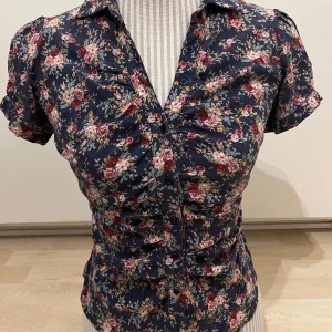 Mörkblå blommig blus med knappar - Säljer en mörkblå blus med korta ärmar och blommigt mönster i rosa, rött och vitt. Blusen har knappar framtill och en figurnära passform med rynkade detaljer. Står S men skulle säga mer Xs. 