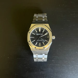 Audemars Piguet klocka - Säljer en Audemars Piguet Royal Oak klocka i silver med svart urtavla. Klockan är i rostfritt stål och har ett automatiskt urverk. Perfekt för den som vill ha en tidlös och stilren accessoar. Klocka är i bra skick, varan skickas samma dag!