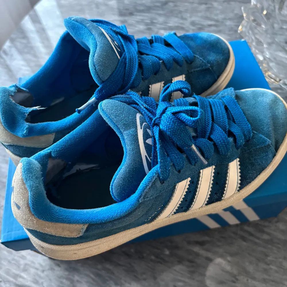 Snygga blå Adidas Campus sneakers med vita detaljer och klassiska tre ränder på sidan. Skorna har en bekväm passform och är perfekta för en avslappnad stil. De är tillverkade i mocka och har snörning framtill.. Kengät.