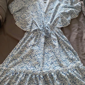 Blommig klänning med volang - Söt blommig klänning i ljusblått och vitt med volangdetaljer. Klänningen har en v-ringad design och ett knytband i midjan för en smickrande passform. Perfekt för sommarens alla tillfällen!