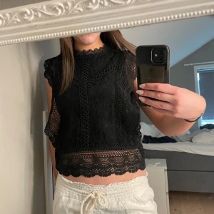 Svart spetsblus - Snygg svart spetsblus med hög hals och ärmlös design. Blusen har vackra spetsdetaljer och en croppad passform som ger en elegant look. Perfekt för att lyfta vilken outfit som helst.