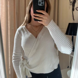 Vit ribbad omlottopp från NA-KD - Säljer en stilren vit ribbad omlottopp från NA-KD. Toppen har en v-ringad design med långa ärmar och en knytning i sidan. Tröjan är i storlek XS men passar mig som är S.  Den är använd fåtal gånger där med väldigt bra skick!🩷