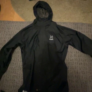 Svart regnjacka från Haglöfs - Säljer en svart regnjacka från Haglöfs med Gore-Tex-material. Jackan har en huva och långa ärmar, perfekt för regniga dagar. 164 cm
