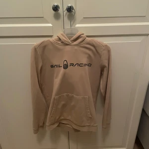 Beige hoodie från Sail Racing - Säljer en beige hoodie från Sail Racing med svart logga på bröstet och ärmen. Tröjan har en klassisk känguruficka och en bekväm huva. Perfekt för en avslappnad stil.
