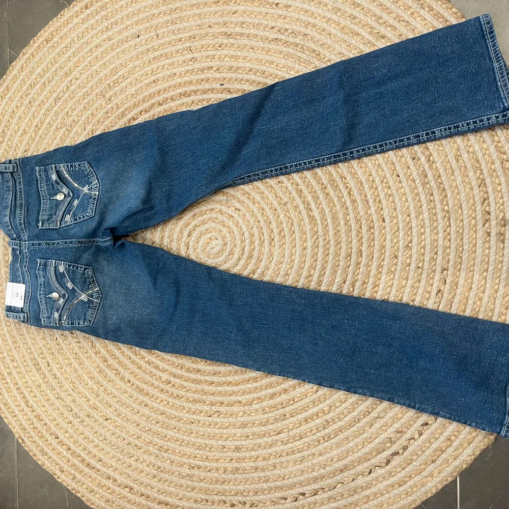 Snygga blå bootcut jeans med dekorativa sömmar och knappar på bakfickorna. Jeansen har en klassisk femficksdesign och en dragkedja framtill. Perfekta för en avslappnad stil.. Farkut & Housut.