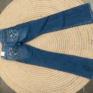 Blå bootcut jeans med detaljer - Snygga blå bootcut jeans med dekorativa sömmar och knappar på bakfickorna. Jeansen har en klassisk femficksdesign och en dragkedja framtill. Perfekta för en avslappnad stil.