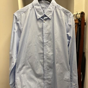 Ljusblå skjorta från Smog - Säljer en stilren ljusblå skjorta från Smog. Skjortan har en klassisk krage och knappar framtill. Perfekt för en snygg och enkel look.