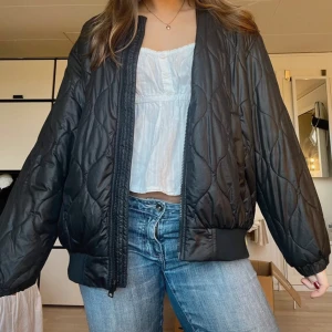 Svart quiltad bomberjacka - Snygg svart bomberjacka med quiltat mönster och dragkedja framtill. Perfekt för en cool och avslappnad stil. Jackan har långa ärmar och ribbade muddar vid ärmslut och nederkant.