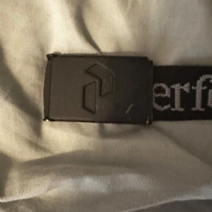 Svart bälte från Peak Performance - Snyggt svart bälte från Peak Performance med vit text längs bandet. Bältet har ett stilrent spänne med logotyp. Perfekt för att ge en sportig touch till din outfit.