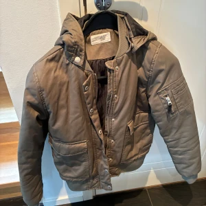 Grönbrun bomberjacka med huva - Säljer en brun bomberjacka med huva och dragkedja. Jackan har knappar framtill och flera praktiska fickor, inklusive en på ärmen. Perfekt för kyligare dagar.