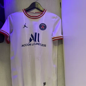 Vit fotbollströja från Jordan x PSG - Snygg vit fotbollströja från Jordan x PSG med korta ärmar och röd-svarta detaljer. Tröjan har PSG-loggan på bröstet och Jordan-märket. Perfekt för fotbollsfans som vill ha stil både på och utanför planen.