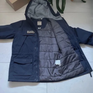 Mörkblå jacka från Napapijri - Säljer en mörkblå jacka från Napapijri med grått foder och huva. Jackan har en norsk flagga på framsidan och flera praktiska fickor. Perfekt för kyliga dagar. MVH EliteVolt