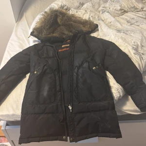 Svart dunjacka från Parajumpers - Säljer en svart dunjacka från Parajumpers med pälskantad huva och dragkedja. Jackan har flera fickor och är en del av Masterpiece Series. Perfekt för kalla dagar!