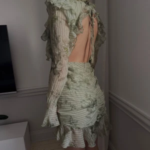 Grön klänning med volanger - Säljer en vacker grön klänning med blommigt mönster och volangdetaljer. Klänningen har en öppen rygg och långa ärmar med volanger vid ärmsluten. Perfekt för en somrig look! Nyskick har lapparna kvar 