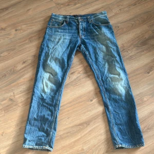 Blå jeans från Nudie Jeans - Säljer ett par klassiska blå jeans från Nudie Jeans. De har en straight passform och är tillverkade i slitstarkt denim. Perfekta för en avslappnad stil. Jeansen har fem fickor och en knappgylf.