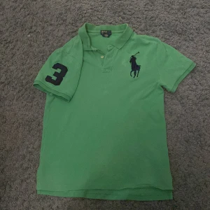 Grön pikétröja från Ralph Lauren - Säljer en snygg grön pikétröja från Ralph Lauren med klassisk krage och knappar. Tröjan har en stor logga på bröstet och siffran 3 på ärmen.