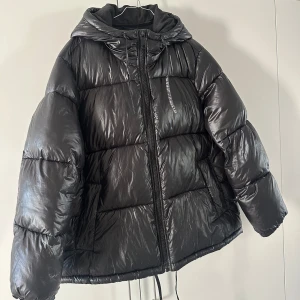 Svart pufferjacka med huva - Säljer en svart pufferjacka med huva och dragkedja. Jackan har en glansig yta och är perfekt för kyliga dagar. Den har långa ärmar och en bekväm passform.